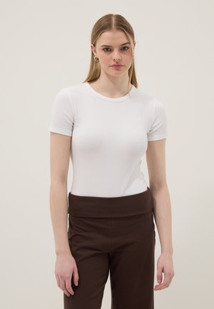 Junge Frau mit langen blonden Haaren, die ein eng anliegendes weißes kurzärmeliges Oberteil und hoch taillierte braune Hosen vor einem schlichten Hintergrund trägt.