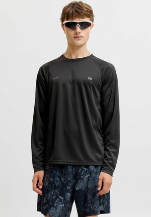 Jeune homme portant une chemise noire à manches longues, un short bleu foncé à motifs et des lunettes de soleil blanches sur un fond clair uni.