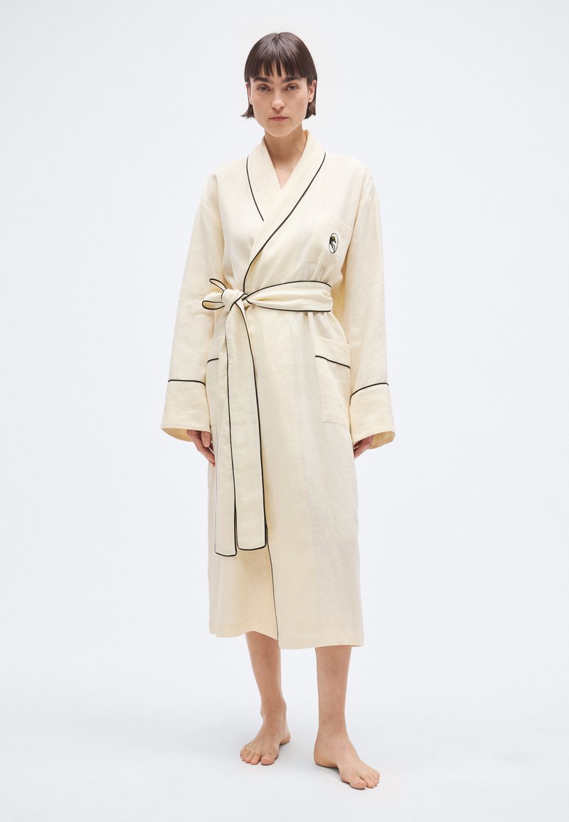 Krēmkrāsas halāts ar melnām apmalēm, garām piedurknēm, sasietu jostu un kabatu kreisajā pusē. Gluda auduma tekstūra.