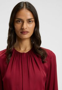 Blusa bordeaux con dettagli plissettati sul colletto e maniche lunghe, realizzata in tessuto leggero che mostra una consistenza liscia.