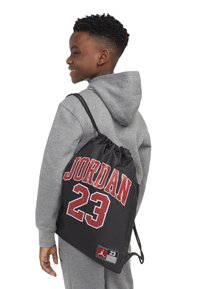 Fekete húzózsinóros táska, amelyen vörös, vastag betűkkel szerepel a "JORDAN" és a "23" felirat. A felülete texturált, és egy oldalsó logófolt található rajta. Szürke kapucnis felsőn viselik.