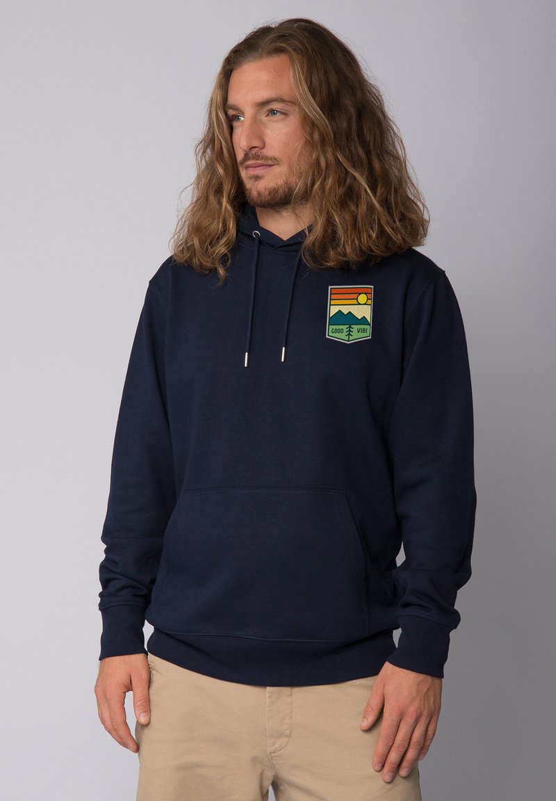 Hoodie bleu marine foncé avec une poche kangourou, un capuchon à cordon de serrage, et un patch graphique représentant des montagnes et un soleil dans des couleurs vives.