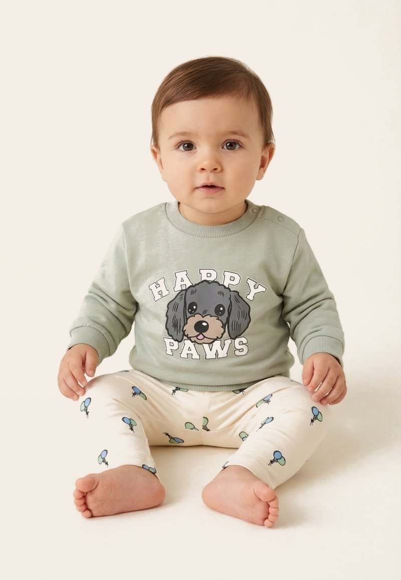 Bébé assis pieds nus portant un sweat-shirt vert clair avec le texte « Happy Paws » et un motif chien, et un pantalon crème avec des motifs de ballons bleus et verts.