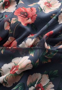 Blåt stof med tekstureret overflade og store røde, hvide og lyserøde blomsterprint med grønne blade, draperet og viser folder og syninger.