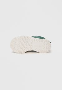 Suola verde di sneaker con gomma bianca testurizzata, caratterizzata da un motivo a zigzag. Il materiale appare resistente e progettato per garantire aderenza.