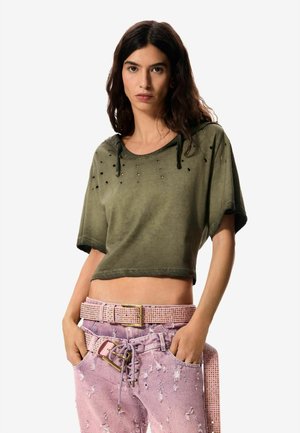 Pikkade tumedate juustega naine kannab oliivrohelist katkendlikku lühikest toppi ja roosaid rebenditega teksapükse, millel on sobiv naelutatud vöö, käed taskutes.