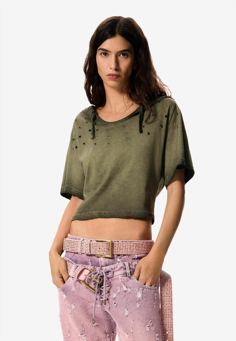 Femme aux longs cheveux foncés portant un haut court kaki déchiré et un jean rose déchiré avec une ceinture cloutée assortie, les mains dans les poches.