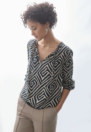 Schwarz-cremefarbene Bluse mit langen Ärmeln, geometrischem Muster, V-Ausschnitt und Kordelzugdetails, kombiniert mit beigen Hosen.
