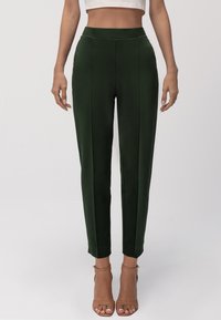 Pantaloni tapered verde satinati con vita alta, caratterizzati da cuciture frontali e una superficie liscia. Abbinati a tacchi a spillo marroni con cinturini.