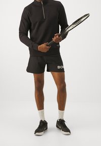 Hombre vestido con ropa deportiva negra y calcetines blancos sosteniendo una raqueta de tenis, de pie contra un fondo claro y liso.