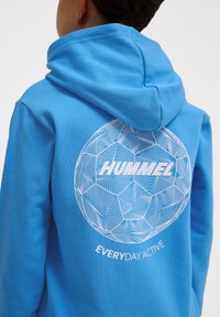 Blaue Kapuzenjacke mit strukturierter Oberfläche, die ein rundes weißes Grafikdesign mit "HUMMEL" und "EVERYDAY ACTIVE" zeigt, das einem stilisierten Fußball ähnelt.