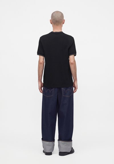 Άνδρας με ξυρισμένο κεφάλι που φοράει μαύρο κοντομάνικο πουκάμισο και σκούρα μπλε φαρδιά τζιν με μεγάλες μανσέτες και μαύρα παπούτσια, στέκεται με την πλάτη προς τα εμπρός.