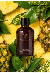 Braune Glasflasche mit schwarzem Deckel, gekennzeichnet mit "True Botanicals Microbiome Enzyme Essence", umgeben von Ananas und grünen Blättern.