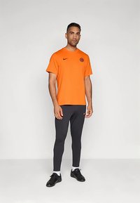 Hombre de pie vistiendo una camiseta naranja de Nike y leggings negros de Nike con calcetines blancos y zapatillas deportivas negras.