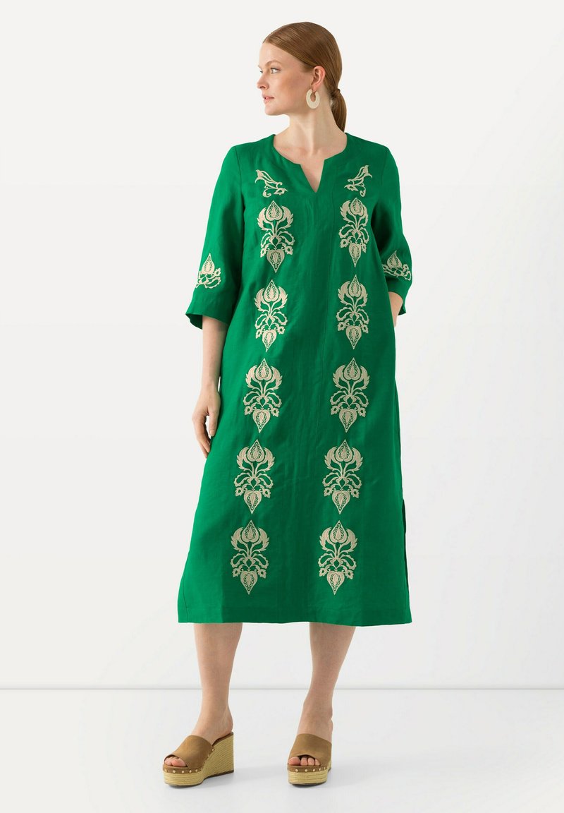Grünes Leinenkleid mit V-Ausschnitt, Dreiviertelärmeln und goldener Blumenstickerei an der Vorderseite. Kombiniert mit beigen Wedgesandalen.