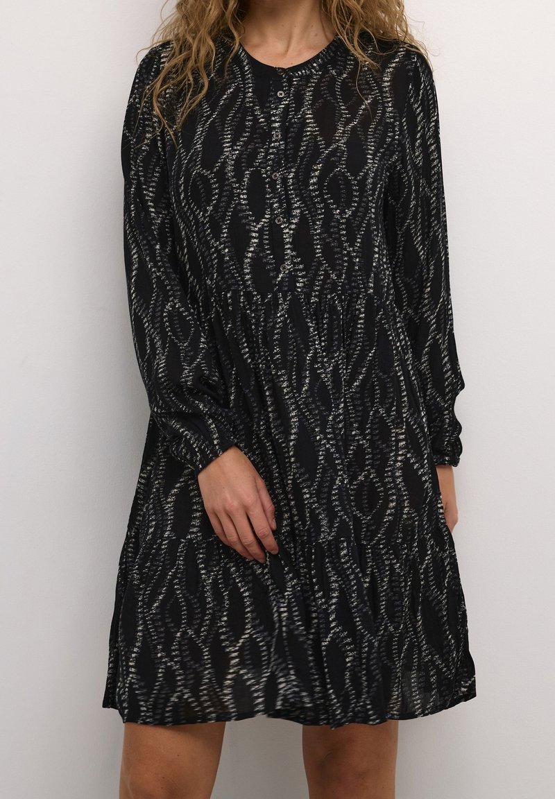 Femme portant une robe noire à manches longues avec un motif abstrait ondulé blanc, des boutons sur la poitrine, debout devant un fond uni.