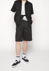 Chemise noire à manches courtes avec boutons et deux poches poitrine, associée à un short noir arrivant aux genoux. Baskets noires, chaussettes blanches avec rayures noires.