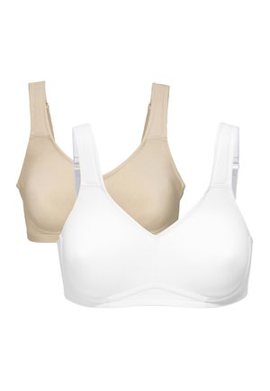 Pitkähiuksinen ruskeatukkainen nainen, joka pitää beigeä langatonta rintaliivit ja korkeavyötäröisiä alushousuja, seisoo valkoisen seinän edessä kasvot eteenpäin.