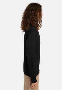 Maglione nero a maglia con collo rotondo, maniche lunghe e polsini a costine, mostrato in vista laterale su uno sfondo bianco semplice.