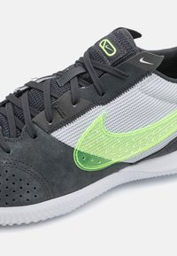 Scarpa sportiva con punta in suede nero e tomaia in mesh grigio chiaro. Presenta un logo verde neon, pannelli testurizzati e una suola in gomma bianca.