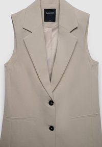 Blazer sans manches beige avec un col cranté, deux poches avant et trois boutons noirs. Intérieur doublé d'un tissu lisse. Étiquette de la marque visible.