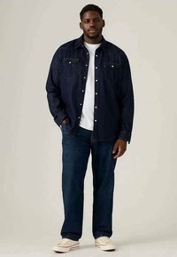 Camisa de ganga escura, com botões, bolsos no peito, usada sobre uma t-shirt branca. Combinada com jeans azul e sapatilhas claras. Ajuste casual.
