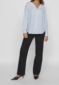 Blouse bleu clair à manches longues avec un col en V et une fermeture à boutons sur le devant, associée à un pantalon noir droit et des talons pointus noirs.