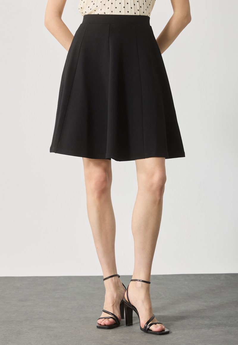 Femme portant une jupe noire évasée longueur genou, un haut beige à pois, et des sandales noires à talons hauts, debout sur un sol gris.