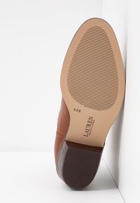 Zapato de cuero marrón con punta redonda y tacón plano. La suela es de color beige claro con líneas de tracción en patrón. Presenta la marca en la parte inferior.