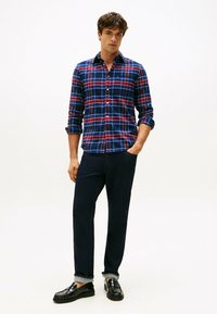 Blau-rotes karogmuster Hemd mit Knopfleiste, kombiniert mit dunklen Hosen und glänzenden schwarzen Schuhen. Modell steht vor einem einfarbigen Hintergrund.