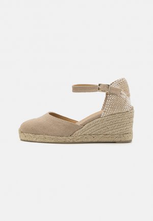 Beige espadrille wedge shoe with a fabric upper, ankle strap, and woven jute sole. The heel features a textured accent panel.