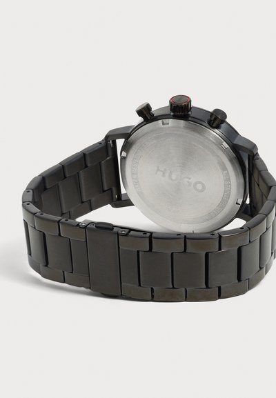 Montre bracelet en métal noir avec dos en acier inoxydable gravé de "Hugo" et trois boutons de contrôle sur le côté.