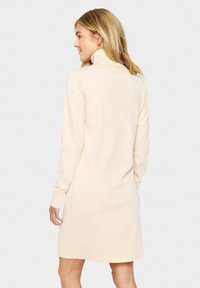 Adīta krēmkrāsas kleita ar augsto apkakli, garām piedurknēm un ribinātu apakšmali; mīksta tekstūra un nedaudz pieguļoša silueta.