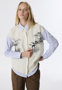 Maglietta a maniche corte con una parte anteriore beige strutturata decorata da illustrazioni botaniche nere, abbinata a una camicia a bottoni blu e bianca a righe sotto.