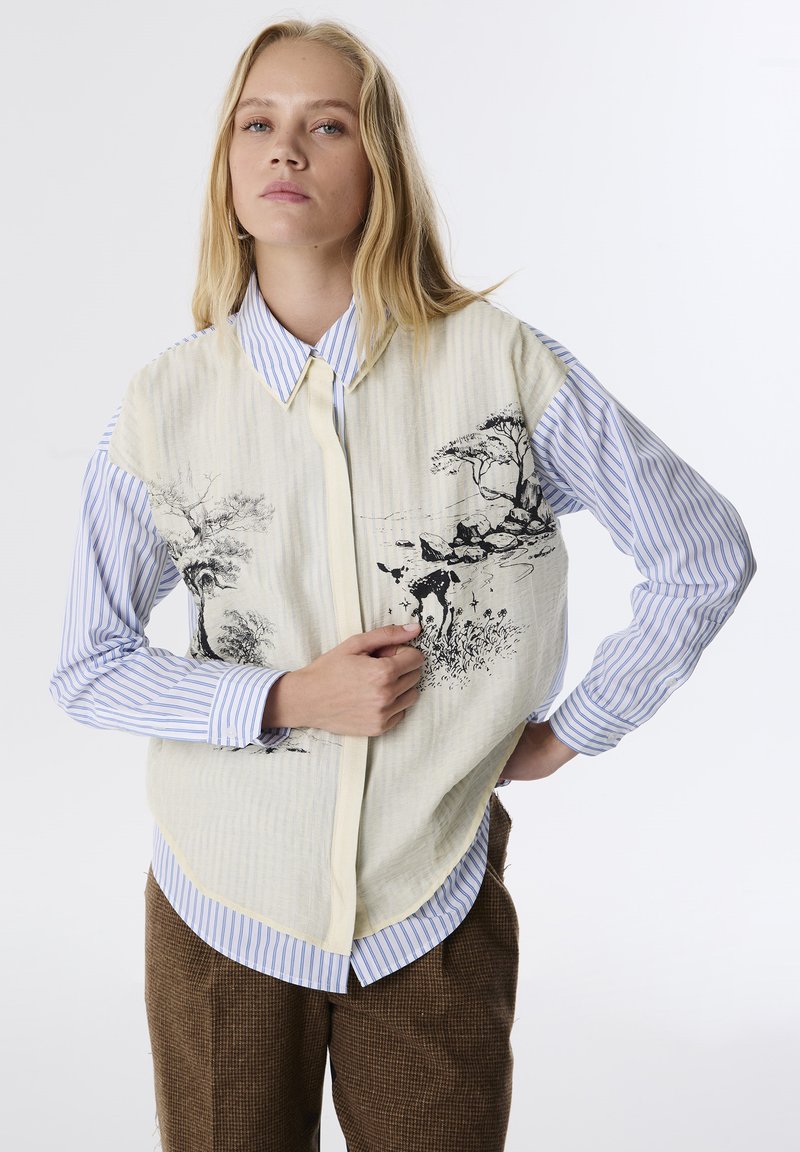 Maglietta a maniche corte con una parte anteriore beige strutturata decorata da illustrazioni botaniche nere, abbinata a una camicia a bottoni blu e bianca a righe sotto.