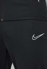 Zwarte sportbroeken van textuurstof, met een rits onderaan en een witte geborduurde Nike-logo op het onderste been.