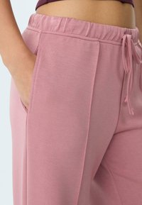 Detailný záber na osobu v ružových teplákoch s elastickým pásom a šnúrkou, pričom jedna ruka spočíva v bočnej vrecku.