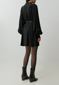 Zwarte jurk met lange, opgeblazen mouwen, tailleriem en een uitlopende rok. Gecombineerd met zwarte enkellaarsjes met hak en doorschijnende panty’s.