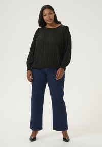 Blouse noire à manches longues avec des rayures verticales et une légère brillance, associée à un jean ample bleu marine et des chaussures noires à bout pointu.