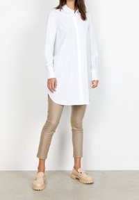 Camicia bianca a maniche lunghe con patta frontale e orlo curvo, abbinata a pantaloni attillati beige e scarpe slip-on di un beige chiaro. Texture morbide in tutto l'outfit.