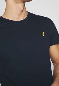 Granatowa bawełniana koszulka z okrągłym dekoltem i krótkimi rękawami, z małym żółtym emblematem ptaka na lewej piersi.