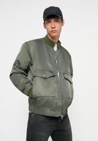 Giacca bomber verde oliva con zip frontale, due ampie tasche a pattina e polsini a costine. Indossata sopra una camicia bianca e abbinata a jeans scuri.