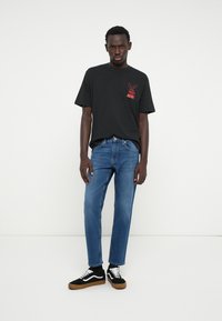 T-shirt de algodão preta com gráfico vermelho, jeans de ganga azul e ténis pretos com detalhes brancos. Modelo está de pé contra um fundo simples.