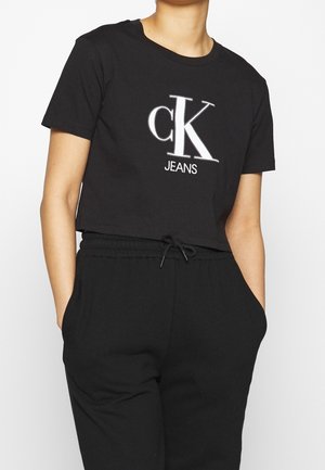 Osoba nosząca czarny krótki t-shirt z dużym białym logo "cK JEANS" oraz czarne spodnie ze ściągaczem, ręce w kieszeniach, jednolite białe tło.