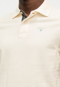 Πουκάμισο πόλο σε εκρού χρώμα με τρία κουμπιά και μικρό λογότυπο Barbour με μπλε ασπίδα στο αριστερό στήθος.