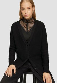 Zwarte gebreide cardigan met een V-hals ontwerp, voorzien van knopen, ribbels aan de mouwen en een gestructureerde stof. Draagt over een zwarte kanten top.