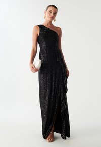 Crna haljina s kristalima za večernje izlaske, s jednim ramenom, nabranim detaljem i visokim prorezom do bedra; uparen s bijelom torbicom i sandalama s trakama.