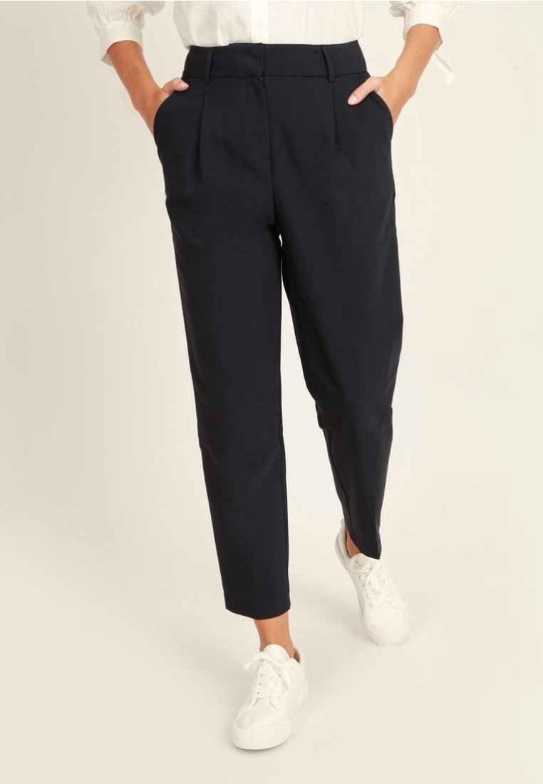 Kvinna som bär svarta högmidjade tapered byxor med veck och vita sneakers, med händerna i fickorna, mot en enkel ljus bakgrund.