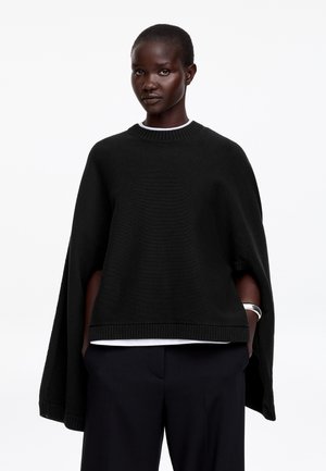 Dark-skinned person wearing a loose black knit poncho over a white shirt and black pants, standing with hands in pockets and a silver bracelet.