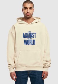 Felpa beige con tasca frontale che mostra il testo blu: "IO CONTRO IL MONDO." Presenta un cappuccio con cordoncino e polsini a coste. Indossata con jeans scuri.